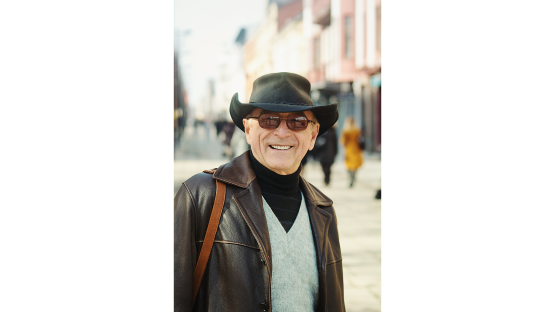  An image of a smiling older man in sunglasses, a v-neck sweater and a cowboy hat. 