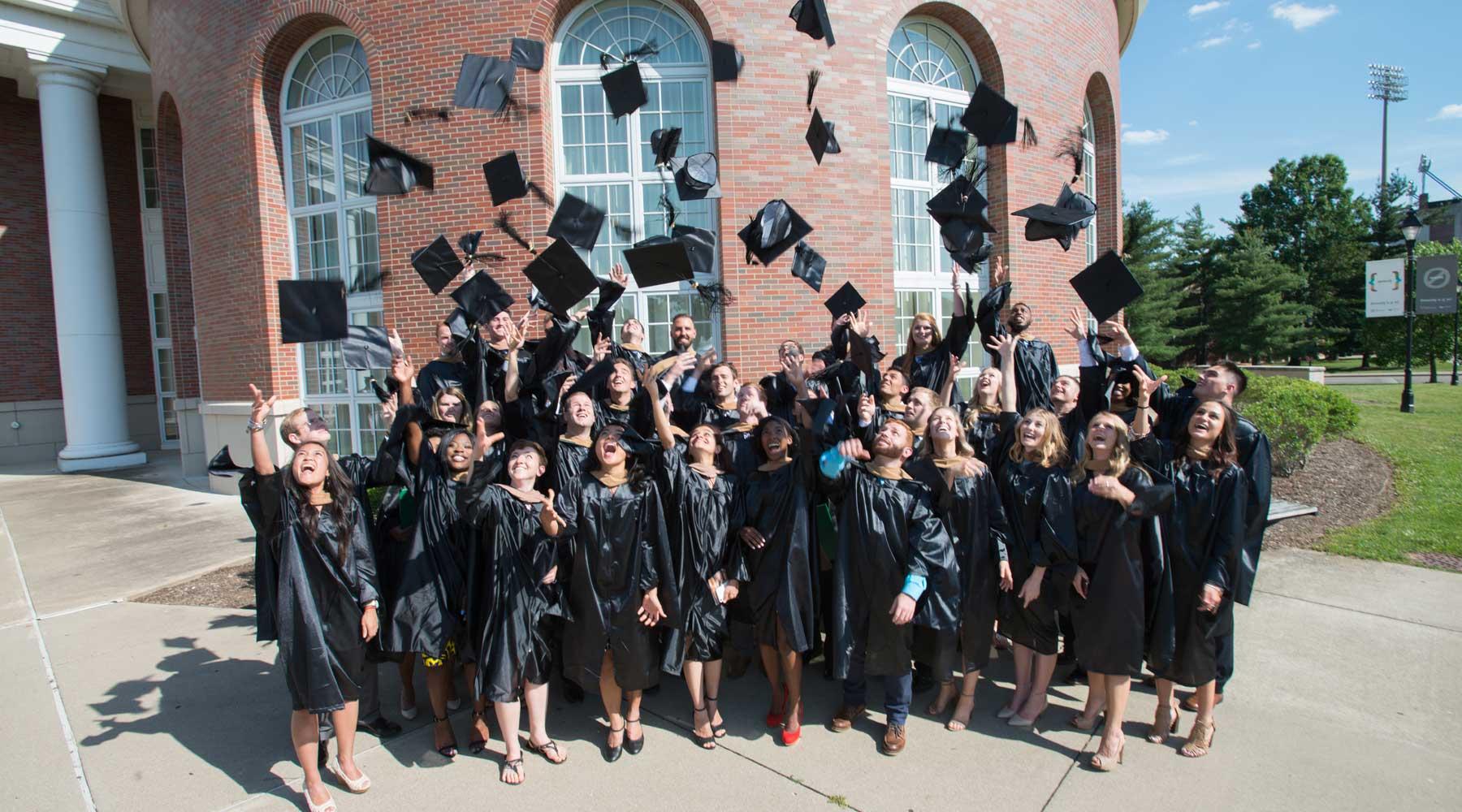 Graduates in black gowns throwing their hats in the air while standing in front of red brick building