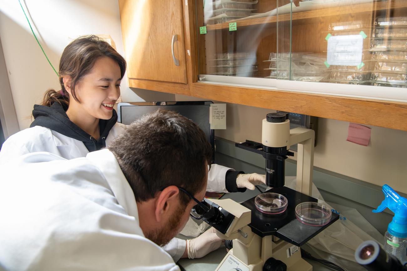 Student smiles as another person in a lab coat looks in a microscope