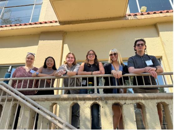 6 students smile and pose over a railing on a building