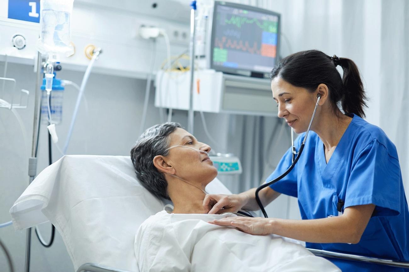 Hospital room with a woman in bed being examined by a student of the online AGACNP program in blue scrubs