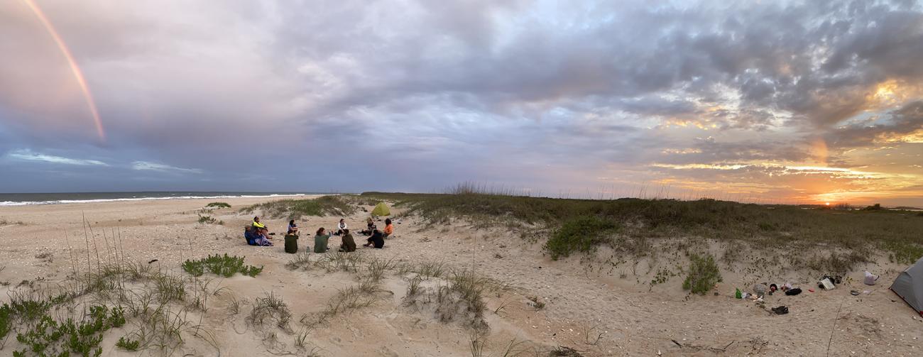 Students sitting on a beach in the distance with a rainbow even further in the distance