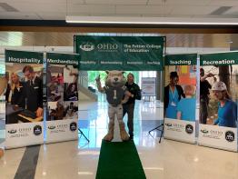 Curt Plummer stands on a green carpet to welcome visitors to Patton Hall surrounded by department banners