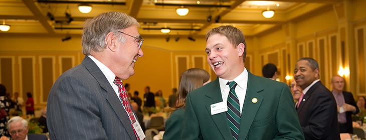 Two professionally dressed gentlemen shaking hands with warm smiles in a banquet room.