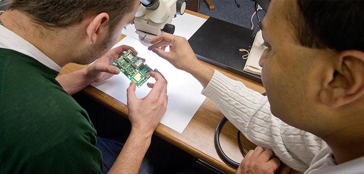 Two men looking at a small circuit. One man is holding the circuit in both hands.