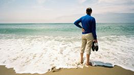 A person wearing a long-sleeve blue shirt and khaki pants standing with hand-on-hip on a beach looking out at the ocean