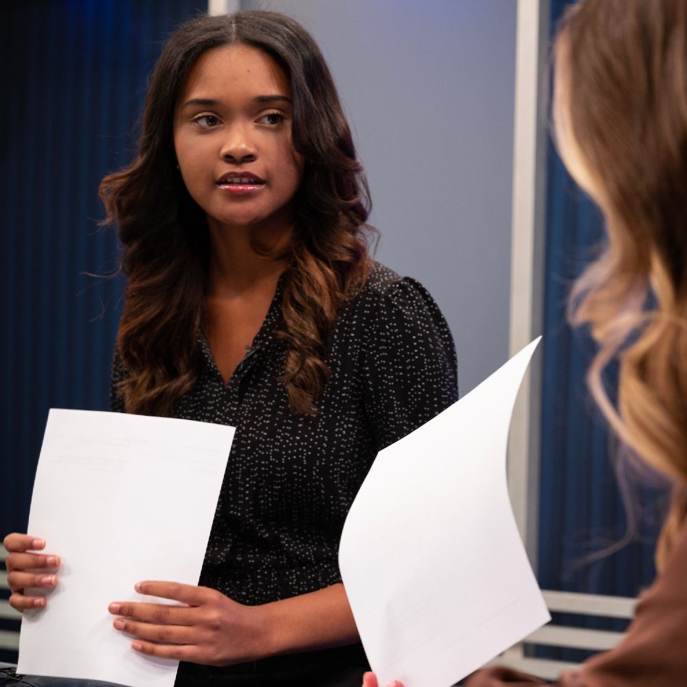 Two women sit and hold papers while speaking with each other