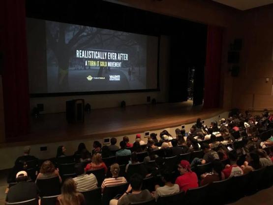 A photo of a cinema with people watching, a video is playing which says 'Realistically Ever After: A Turn it Gold Movement'