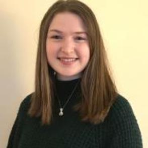 Caroline Chalk stand in front of a cream colored wall, smiling at the camera and wearing a black turtleneck.