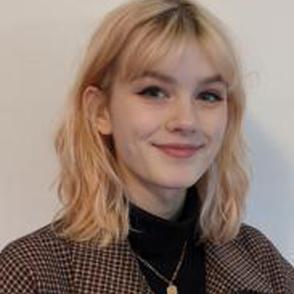 Cora Fitch stands in front of a white wall, she wears a black shirt with a tweed plaid jacket over it.