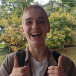 Elena Detach wearing an orange button up over a white shirt, holding two thumbs up with a smile on her face.