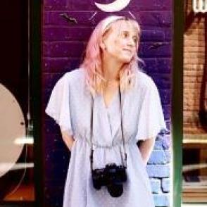 Gwen Ehlers in a blue dress with a camera around her neck, posed in front of a deep purple wall with a crescent moon painted on it.