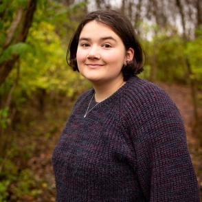 An image of Kate Wiselogel in a forest, wearing a blue-purple sweater.