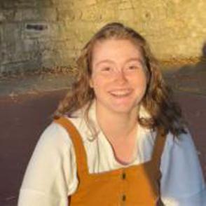 Makenna Koogler smiling in the sun wearing a white long sleeve shirt with yellow corduroy overalls over it.
