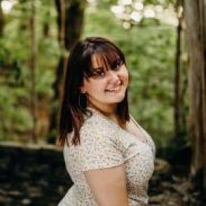 Olivia Mincks standing in a forest, turned towards the camera smiling while wearing a white floral dress.