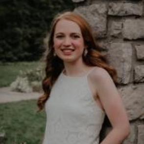 Samantha Schmmoller leans against a rocky post. She has one hand in her pocket, smiling at the camera and wearing a white tank top.