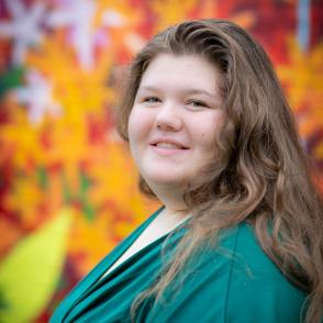 An image of Maggie Giansante in a deep green dress with orange leaves behind her.