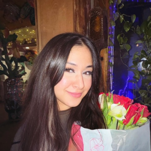 Fizza Stands in front of a yellow wall, a window next to her. She wears a black shirt and carries a bouquet of flowers.