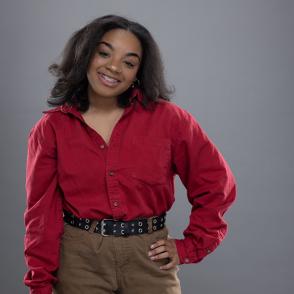 Laila Christian stands in front of a gray wall. She wears a long sleeve red button up with a black belt and brown pants. She has one hand on her hip.