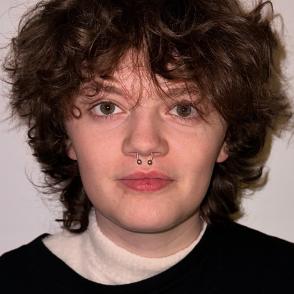 Mallory Just stands in front of a white wall with a neutral expression on her face. She wears a black Led Zeppelin T-shirt over a thin white turtleneck. She also has a silver septum piercing.