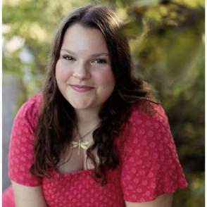 Maya Shaw leans toward the camera and smiles. She wears a red dress that has a lighter dotted pattern. She also wear a gold dragonfly necklace.