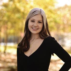 Emily Fassbender stands in front of distant trees, smiling at the camera with one hand on her hip.