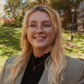 Becky Ivan smiles and wears a ruffled high-collared black blouse and a gray tweed blazer and glasses