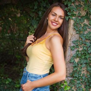 Cassidy Dummitt leans against a wall and smiles, wearing jeans and a light yellow tank top