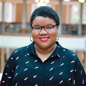 Caylah Awls smiles at the camera, wearing glasses and gold hoops, as well as a navy blue button-up shirt with a reoccurring lipstick tube pattern.