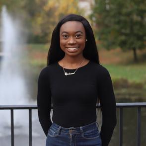 Jennifer Ayarkwa stands in front of a fountain and black railings, smiling and wearing a black long sleeve shirt and jeans.