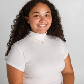 Mia Walsh stands in front of a background, smiling at the camera, wearing a white short-sleeved shirt with a high neck and a gold chain