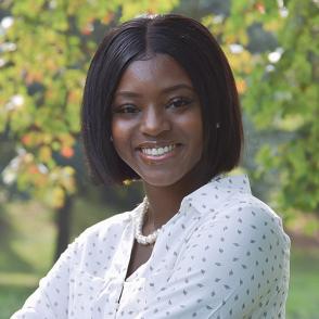 Nia Dumas smiles at the camera outside, wearing a white collared shirt with a small leaf pattern and a white pearl necklace.