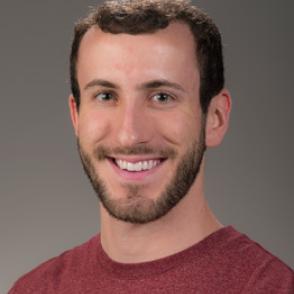 Nicholas Whitticar smiles at the camera wearing a red shirt in front of a gray background