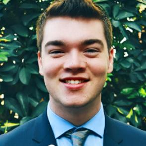 Rhyan Goodman smiles wearing a light blue collared shirt, dark blue blazer, and blue and yellow striped tie