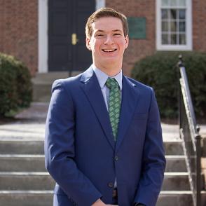 Samuel Carryer wears a navy suit and a light green and white tie, smiling at the camera with his hands in front of him.