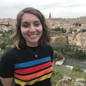 Suzy Aftabizadeh stands with her hands behind her back, smiling at the camera and wearing a retro striped shirt.