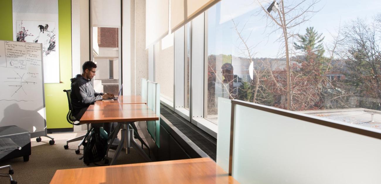 A student uses a laptop by windows overlooking campus.