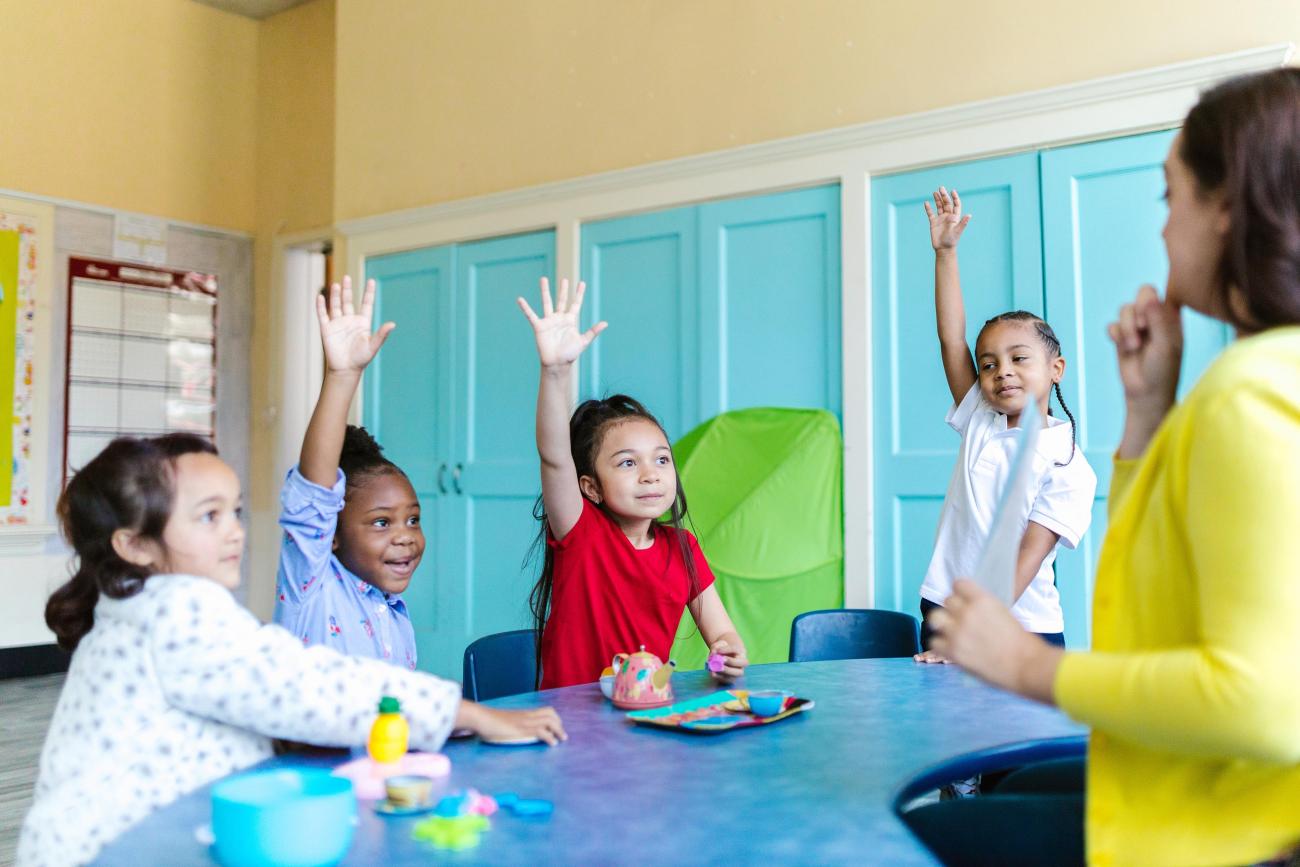 a teacher stands in front of young children with raised hands