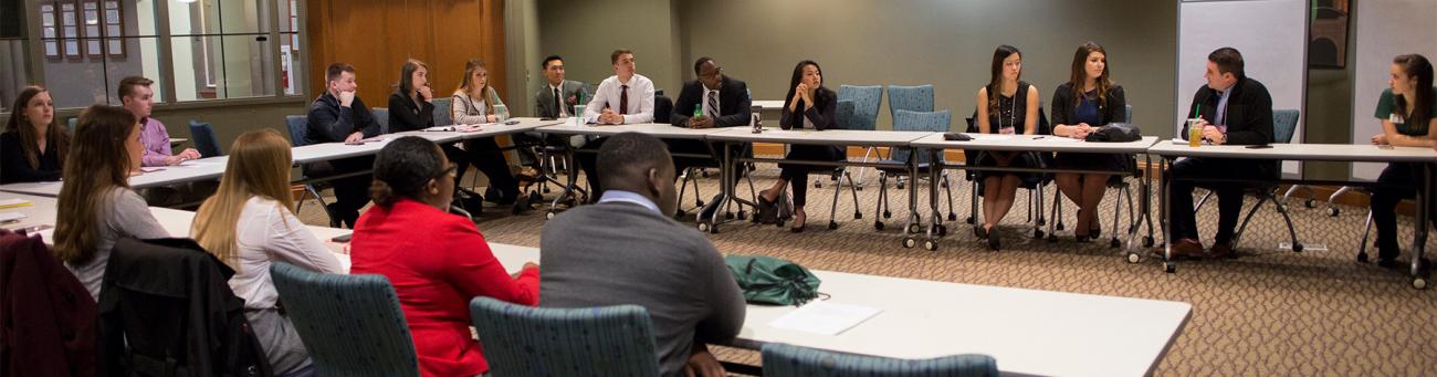 Many people sit at a large U-shaped table in a room having a discussion.