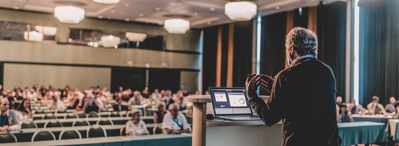 A professor stands in front of a podium while lecturing to a classroom full of people