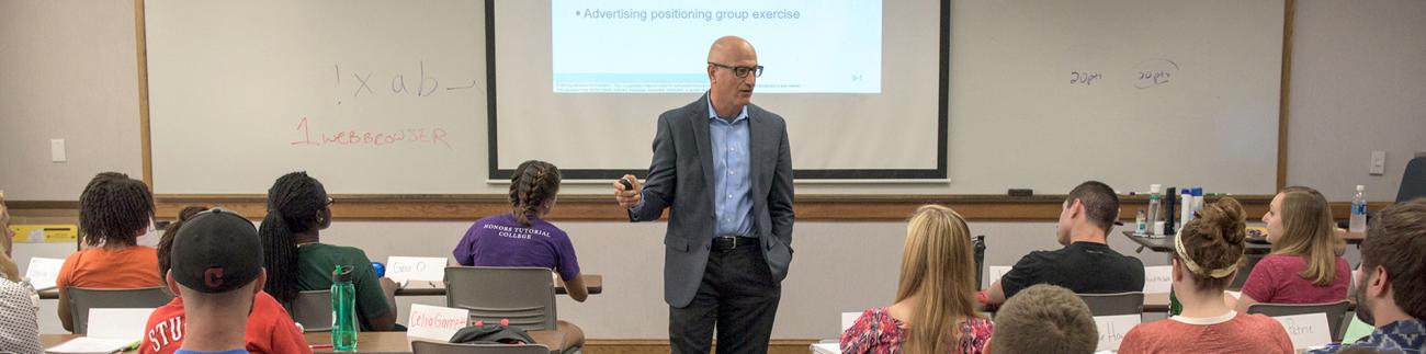 A professor stands in front of a projector screen teaching a class of students.