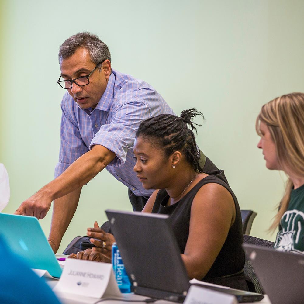 An instructor stands beside a student pointing at their laptop as they explain something.