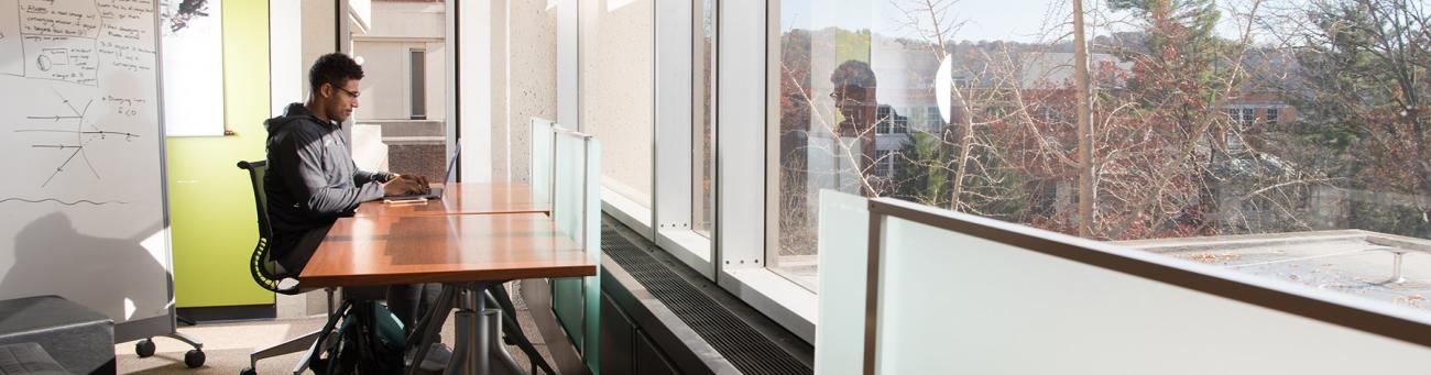 A student sits at a desk in front of a window overlooking campus while working on his laptop.