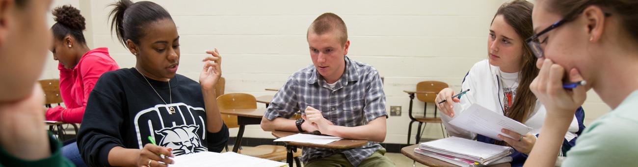 Five students sit with their desks pushed together discussing an assignment.