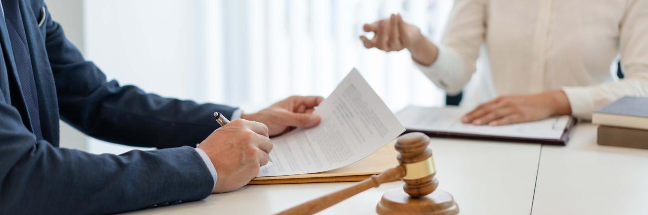 Person in suit with pen above a document with another person in background and a gavel on the table
