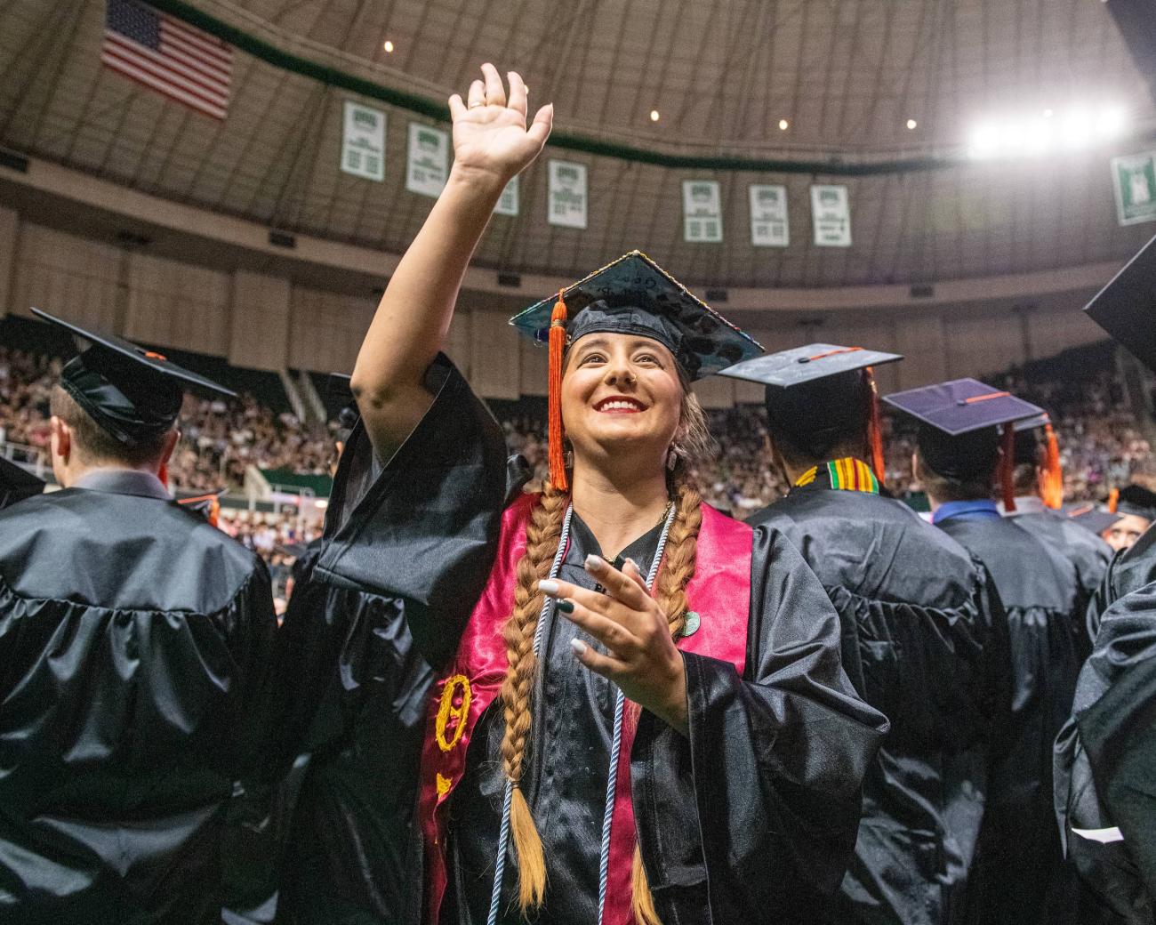 Student smiles and waves wearing their cap and gown at graduation
