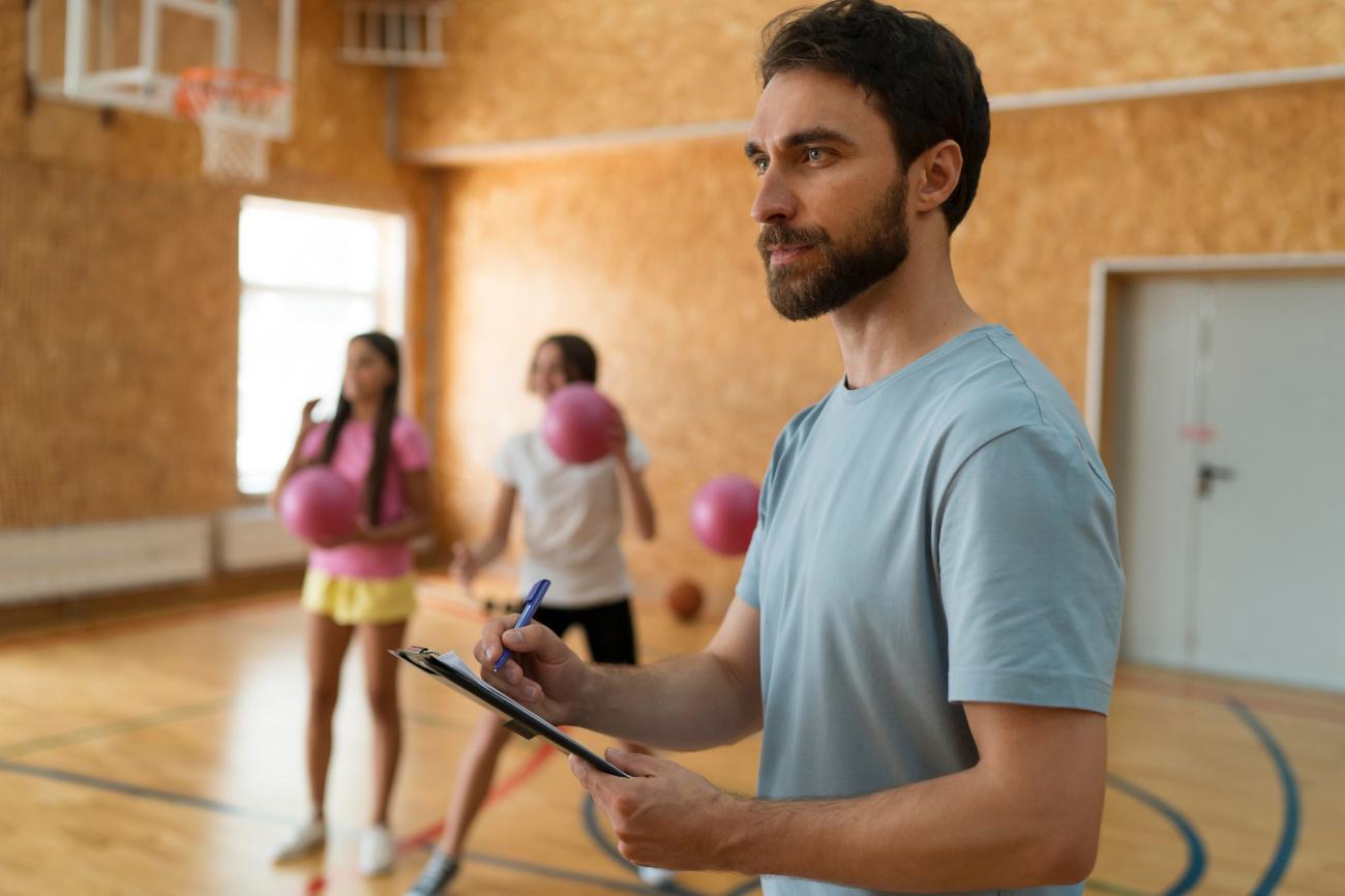 Man writing on clipboard with children in background throwing dodgeballs. Image by Freepik.