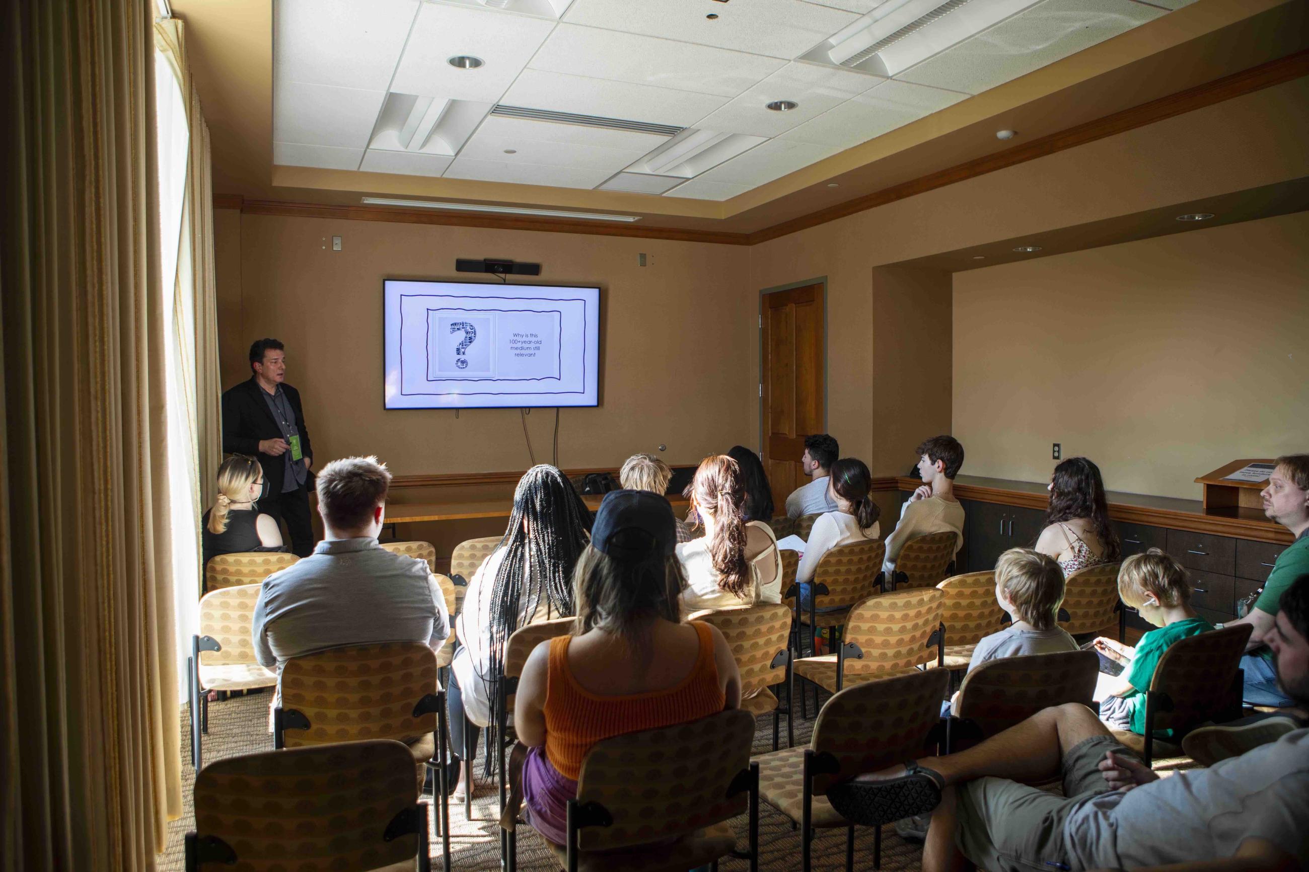 Jon Zellner standing at front of room next to screen presenting to a room of about  20 people