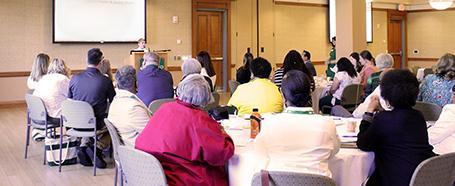 People sit at round tables listening to a presentation in Baker Center room 240-242
