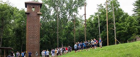 People lined up wearing high ropes gear, next to a climbing wall structure outdoors